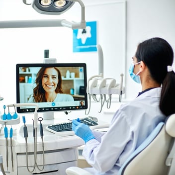 The image depicts a modern dental office equipped for teledentistry. In the foreground, a dentist is seen engaging with a patient via a large screen displaying a video consultation, highlighting advanced technology. The room is well-lit and organized, featuring dental tools neatly arranged on a counter. A digital display shows a virtual waiting room with a welcoming atmosphere. In the background, a poster on the wall promotes remote dental services, emphasizing convenience and accessibility. The dentist, wearing a white coat and protective gear, appears attentive and professional, engaging with the patient who smiles from their home setting. A small logo in the corner represents the practice, integrating themes of innovation and patient-centered care. Overall, the image encapsulates the fusion of traditional dentistry with modern telehealth solutions, showcasing the future of dental care.