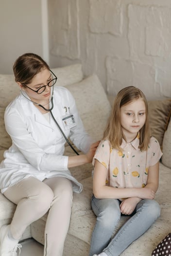 A clinician sits on a couch in a home setting and listens to a young girl's lungs using a stethoscope in the patient's house.